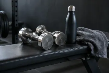 Deux haltères métalliques, une gourde noire et une serviette grise posées sur un banc de gym